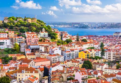 Serene blue water appears in the background, colorful structures with terracotta roofs appear in the foreground. It is a sunny day.